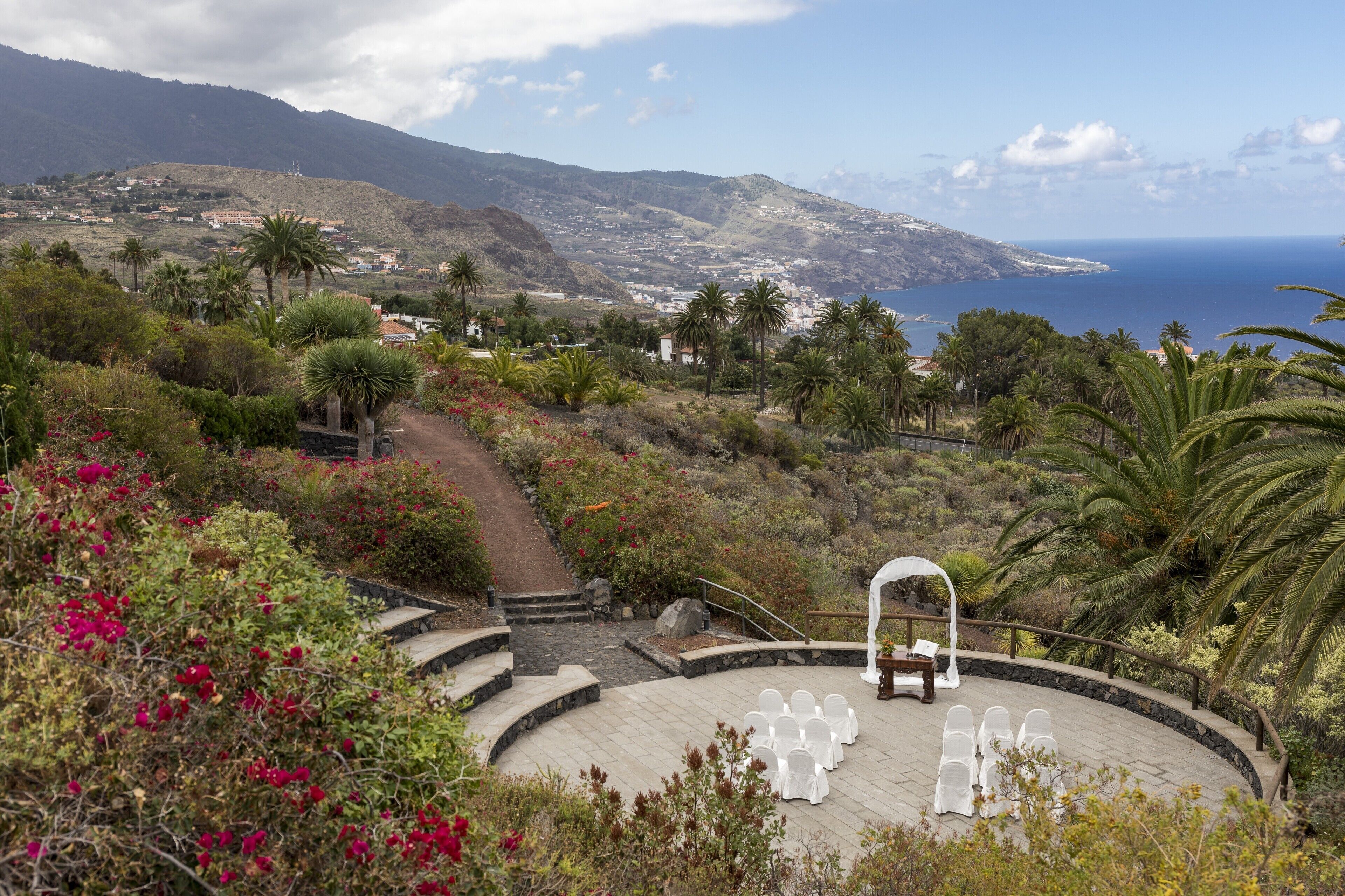 A circular outdoor seating area with white chairs and a wooden bench, surrounded by lush greenery and a view of the sea and mountains.