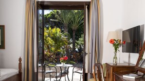 Una habitación con piso de madera, ventilador de techo, cama, escritorio con silla, televisor y vista a un jardín a través de una puerta de vidrio.