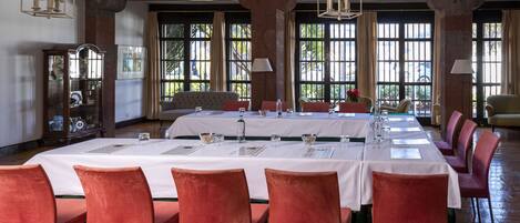 Un comedor con una larga mesa dispuesta para una comida, rodeada de sillas rojas, con amplios ventanales y una araña.