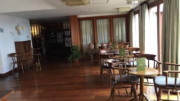Een ruime kamer met houten vloer, een groot raam met gordijnen, en verschillende houten tafels en stoelen.