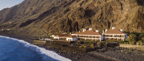 A coastal property with a large house featuring multiple balconies, situated on a rocky beach with a view of the ocean and a mountainous backdrop.
