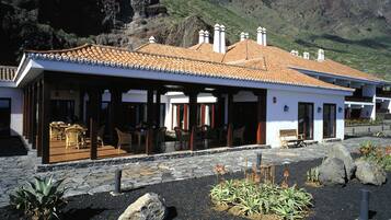 A building with a tiled roof, a covered patio with chairs, and a garden with rocks and plants.