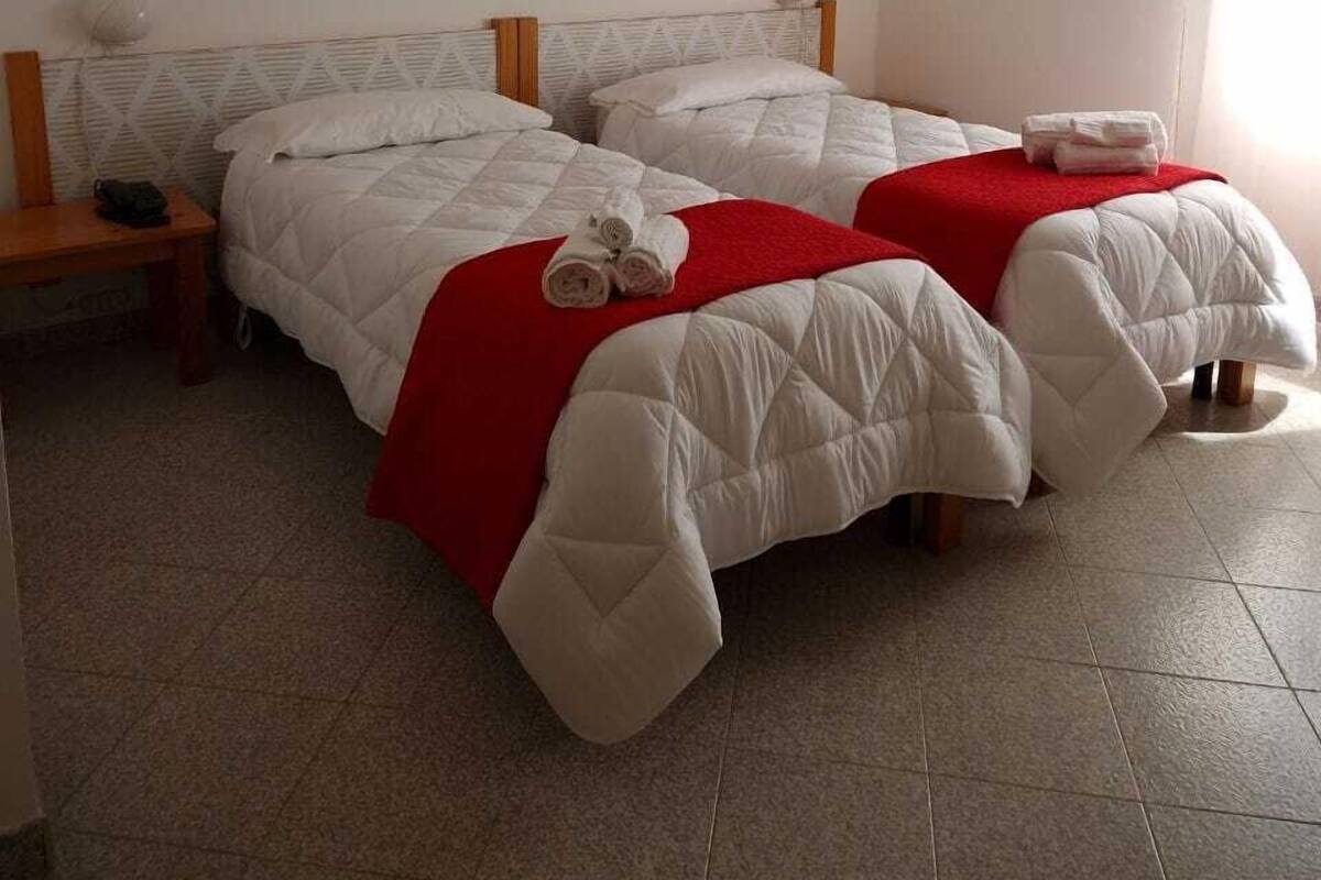 Une chambre avec deux lits, chacun recouvert d’une courtepointe blanche matelassée et d’une couverture rouge. Une décoration murale est accrochée au-dessus des lits, et une petite table avec une lampe se trouve à côté de l’un des lits.
