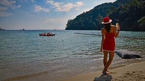 A woman in a red dress and hat standing on a beach, looking out at the sea with boats in the distance. - Eight Hotel Portofino (Portofino)