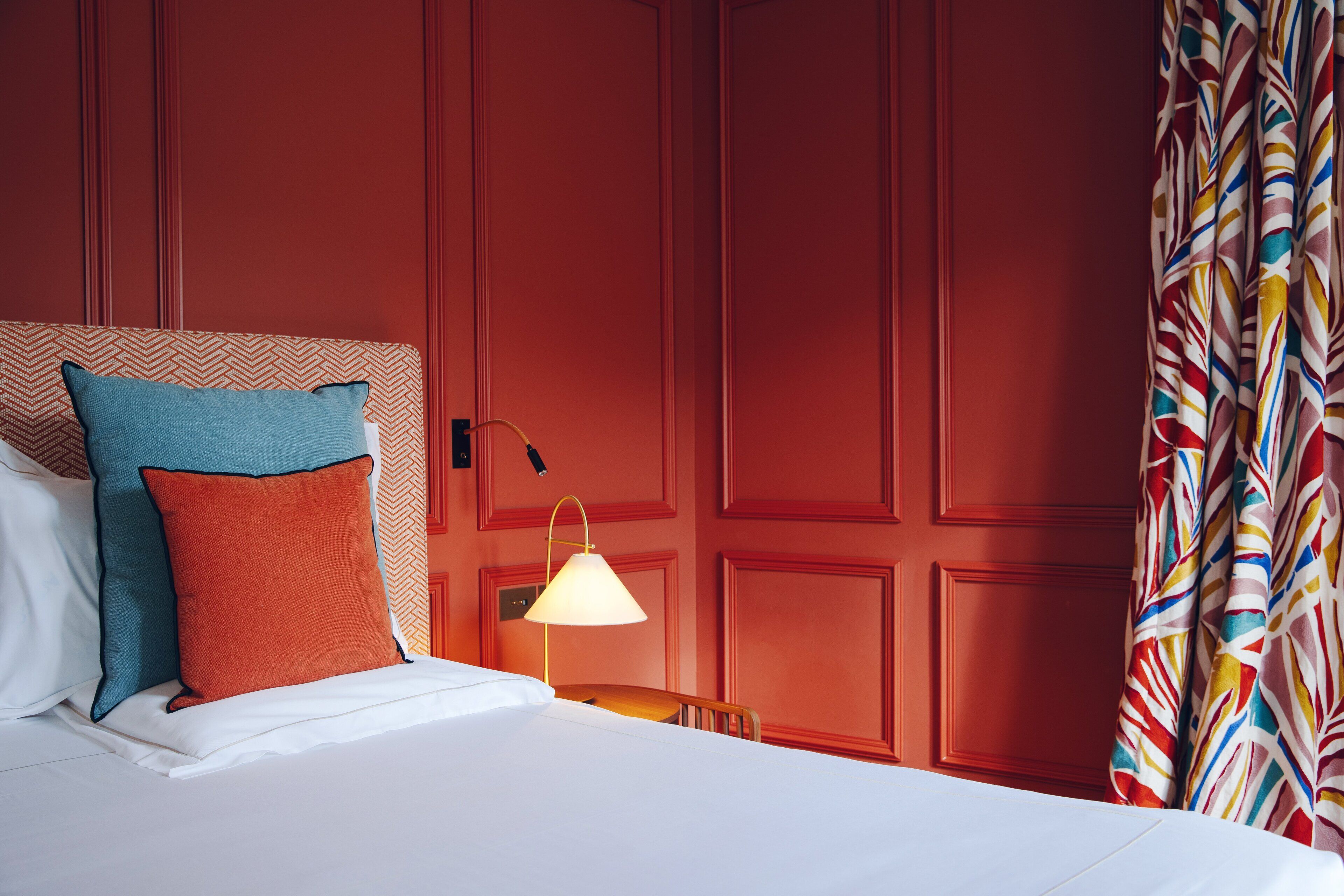 A hotel room with a bed, pillows, a nightstand with a lamp, and a red wall with paneling.