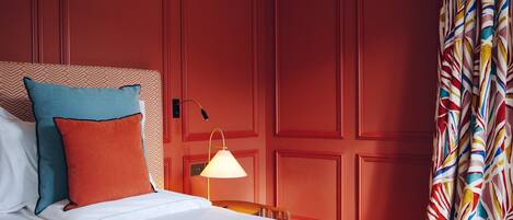 A hotel room with a bed, pillows, a nightstand with a lamp, and a red wall with paneling.