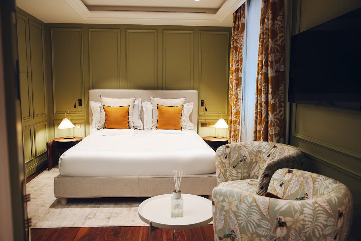 Ein Hotelzimmer mit einem großen Bett, zwei Nachttischen mit Lampen, einem runden Couchtisch und einem Sessel mit Blumenmuster.