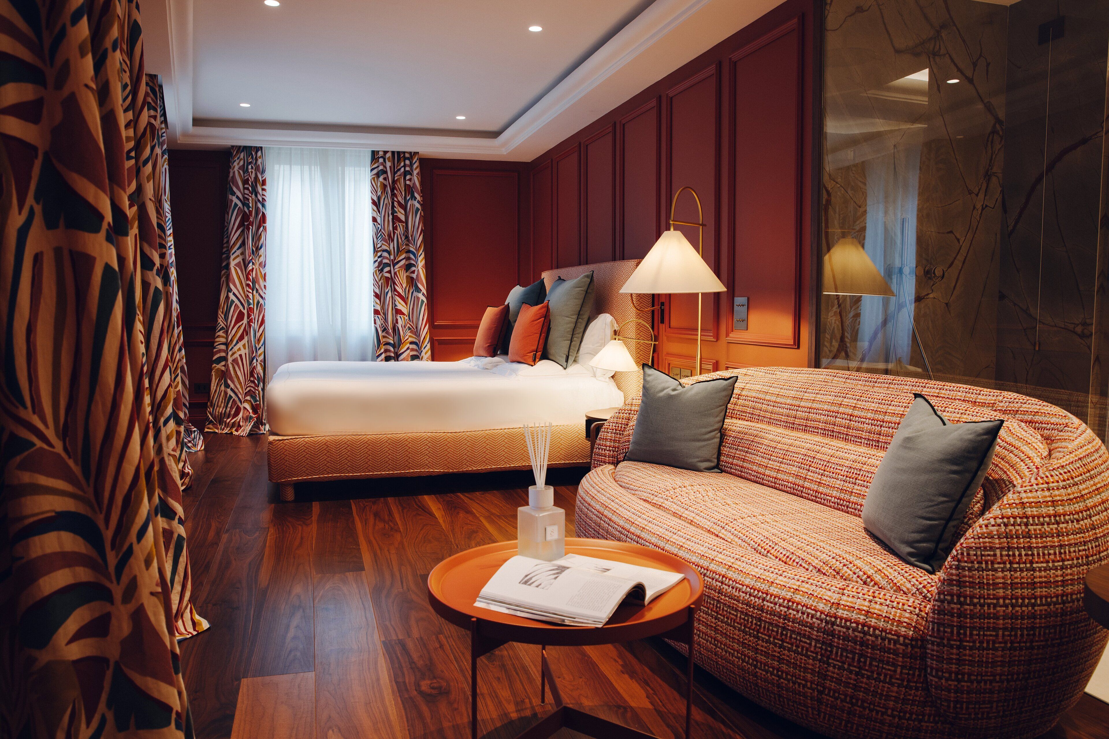 Una camera d'albergo con un letto, un divano, un tavolino rotondo con una rivista e una lampada.
