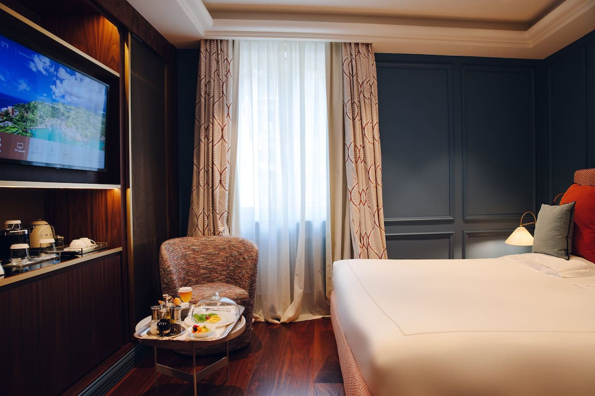 Ein Hotelzimmer mit einem Bett, einem an der Wand befestigten Fernseher, einem Sessel, einem kleinen Tisch mit Frühstücksgebäck und einem Vorhangfenster.
