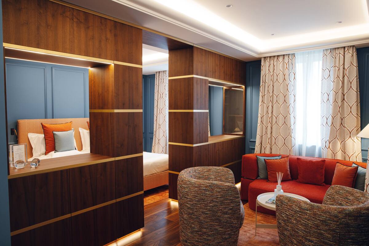 Ein modernes Hotelzimmer mit einer Wand in Holzoptik, einem roten Sofa, einem runden Couchtisch und einem großen Bett mit vielen Kissen.