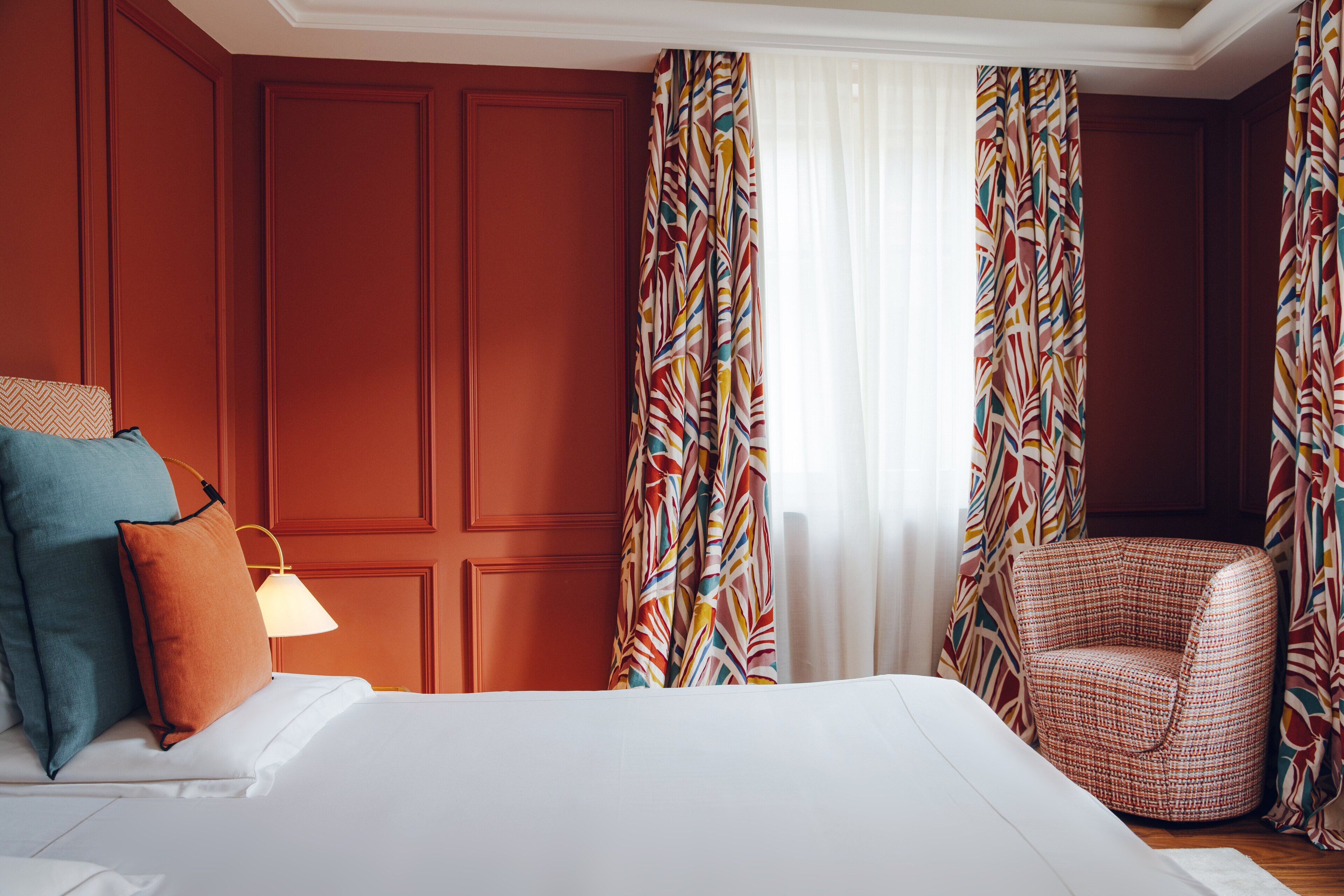 A hotel room with a bed, pillows, a lamp, a chair, and curtains.