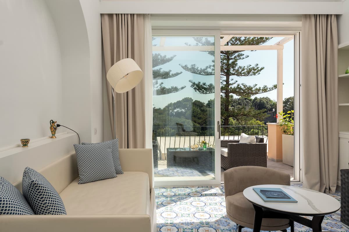 A modern hotel room with a sofa, a small table, and a balcony with a view of trees and a garden.