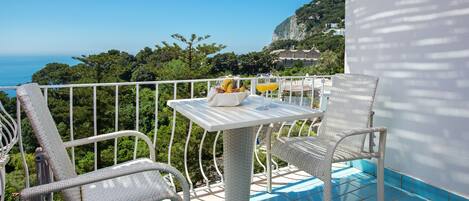 A balcony with wicker furniture, a table with a fruit bowl, and a view of the sea and greenery.