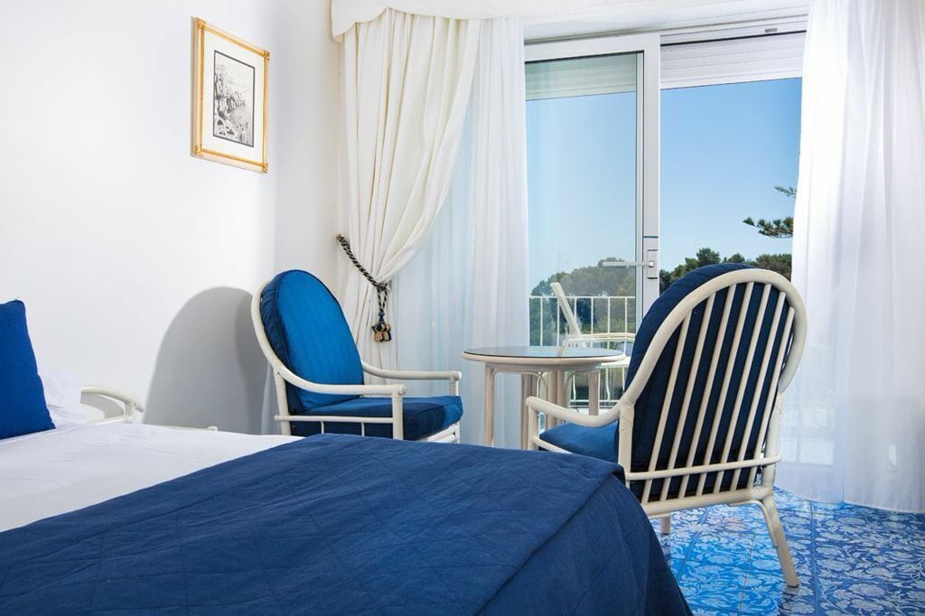 Habitación de hotel con cama, dos sillas, una mesita y un balcón con vistas.