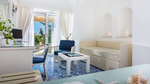 A room with a blue and white tiled floor, a white sofa, a blue chair, a small white table, a TV on a white stand, and a window with curtains. - Hotel La Vega (Capri)