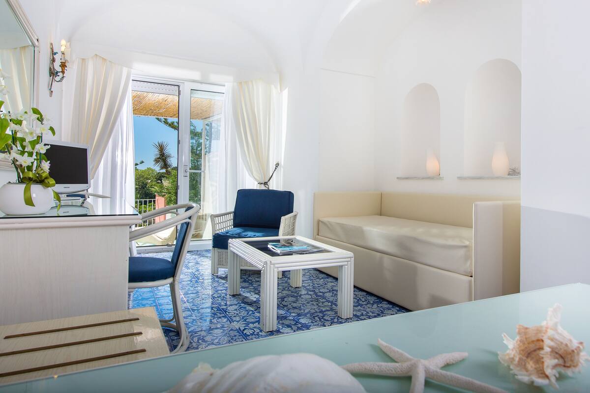 A room with a blue and white tiled floor, a white sofa, a blue chair, a small white table, a TV on a white stand, and a window with curtains.