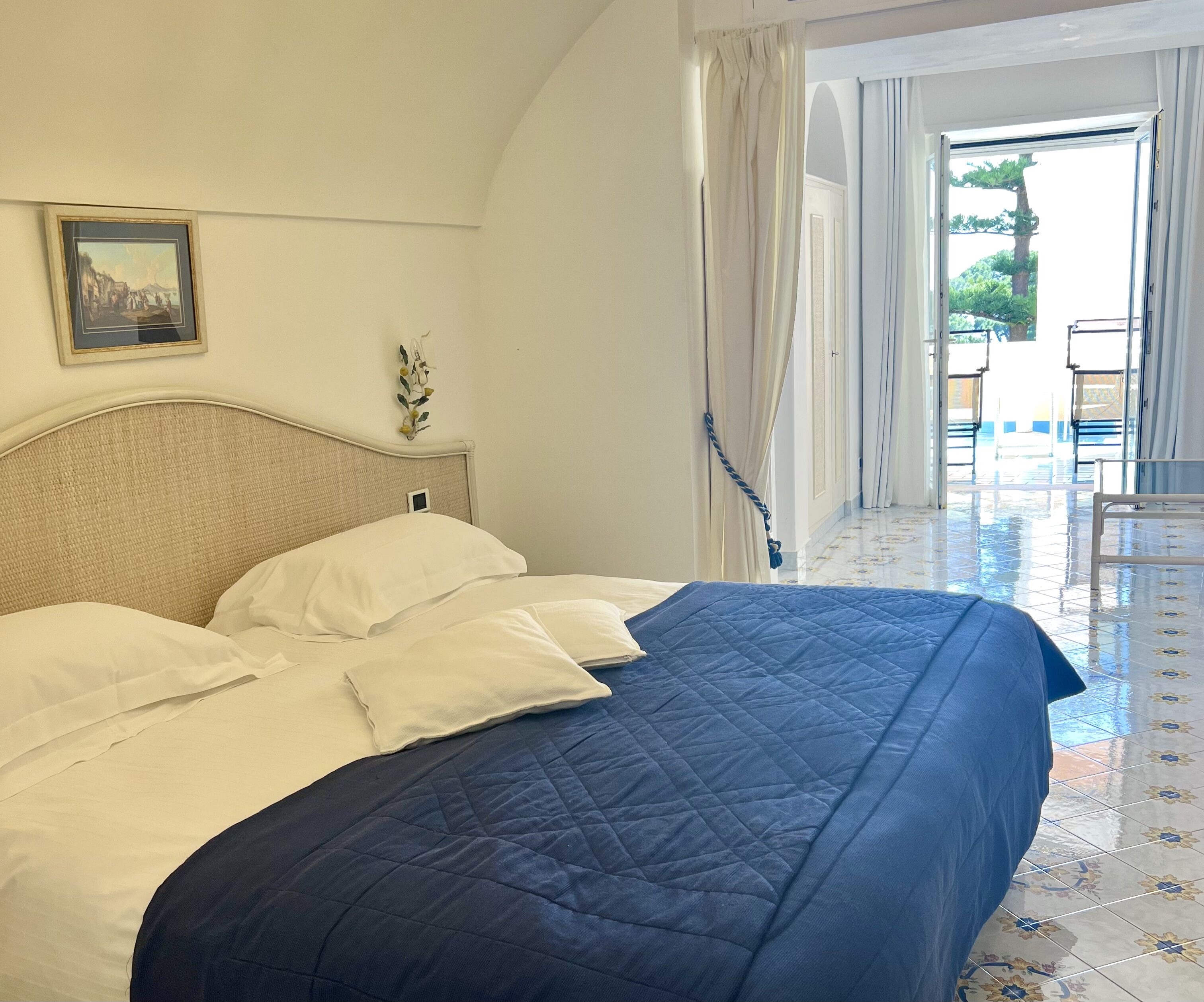 A neatly made bed with white and blue bedding, a framed picture on the wall, and a view of the outdoor area through an open door.