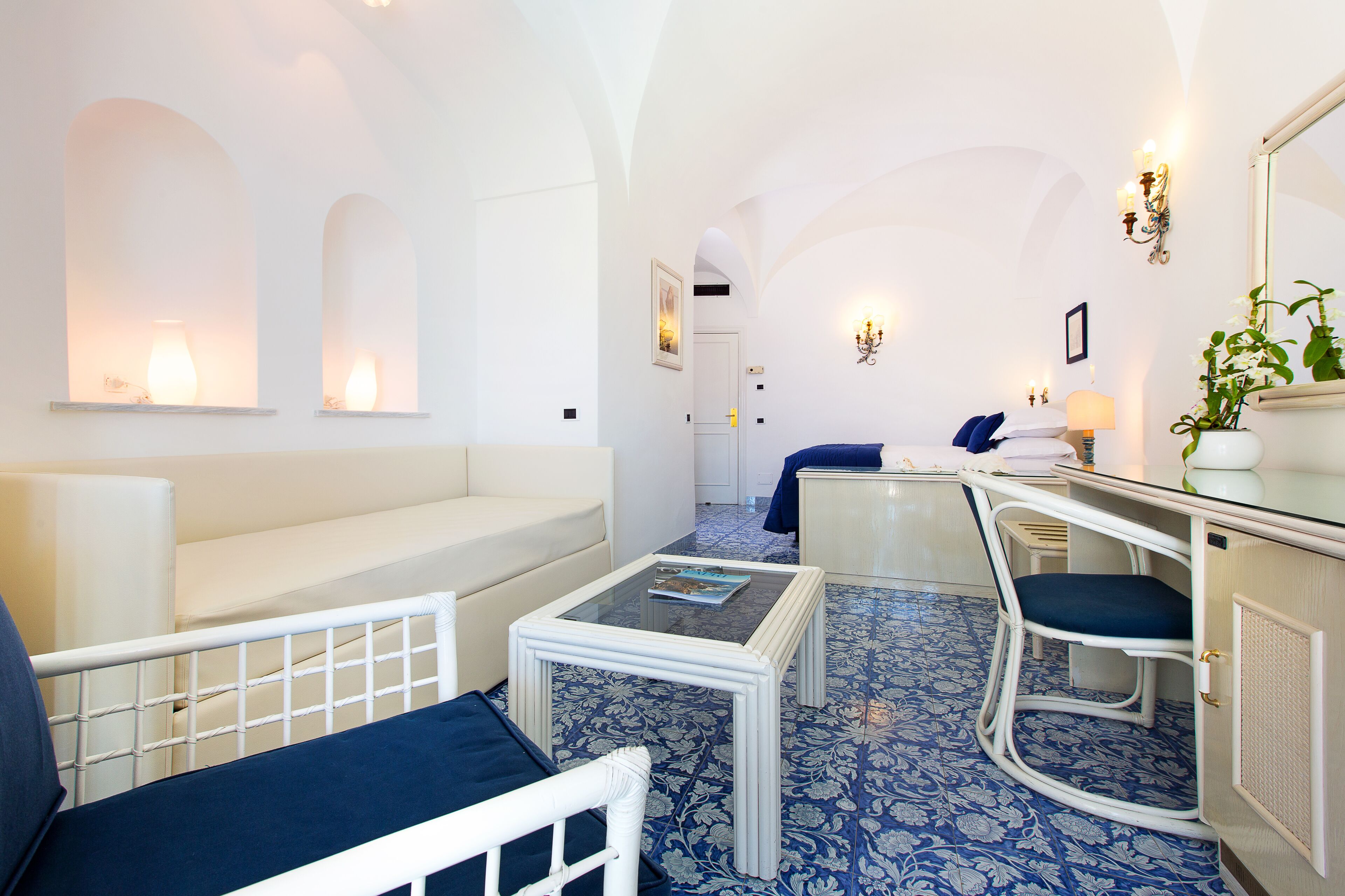 A hotel room with a white sofa, a small table, a desk with a chair, and a bed.