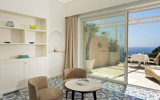 Una habitación con un amplio ventanal corredera que da a un balcón con vistas al mar. Hay dos sillones color beige y una pequeña mesa redonda con un cuenco decorativo. La habitación tiene paredes blancas, suelo de baldosas y un armario blanco con estantes.