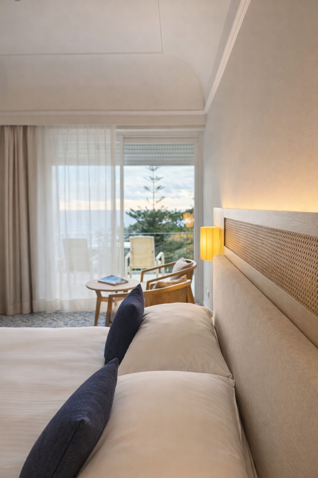 Habitación de hotel con cama, mesita de noche, lámpara, ventana con vista al exterior y una silla junto a la ventana.
