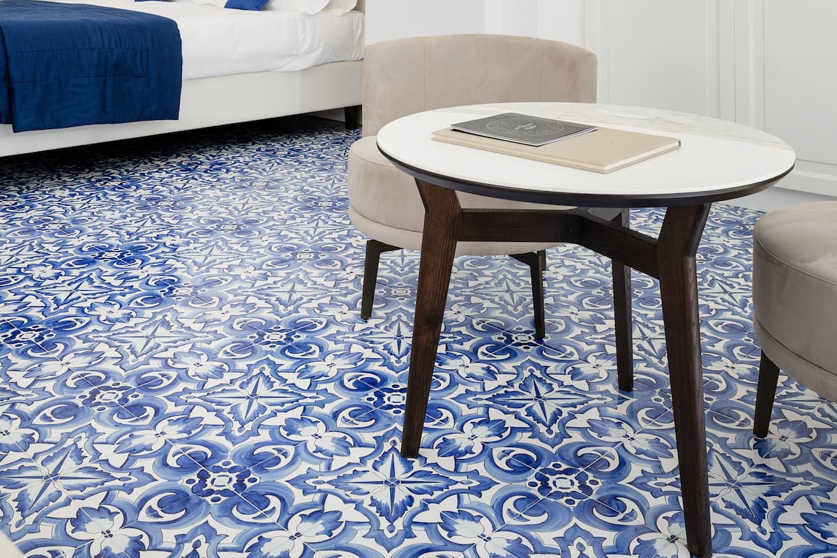 A modern hotel room with a white bed, blue patterned floor, a round table, and a bookshelf.