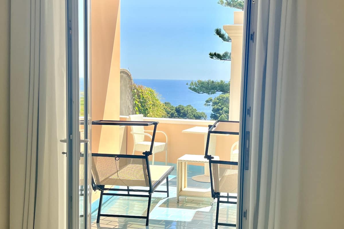 A balcony with a glass door, white curtains, and a view of the sea.
