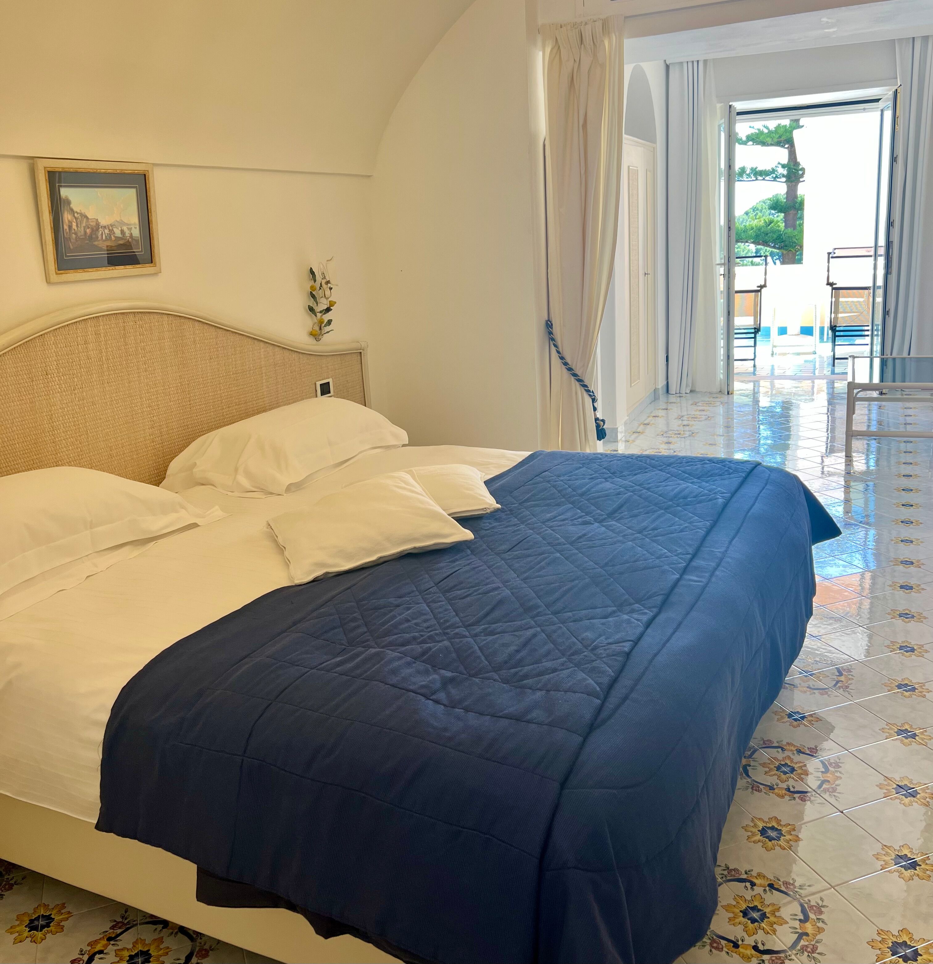 Una cama bien hecha con ropa de cama blanca y azul, un cuadro enmarcado en la pared y vistas del exterior por la puerta abierta.