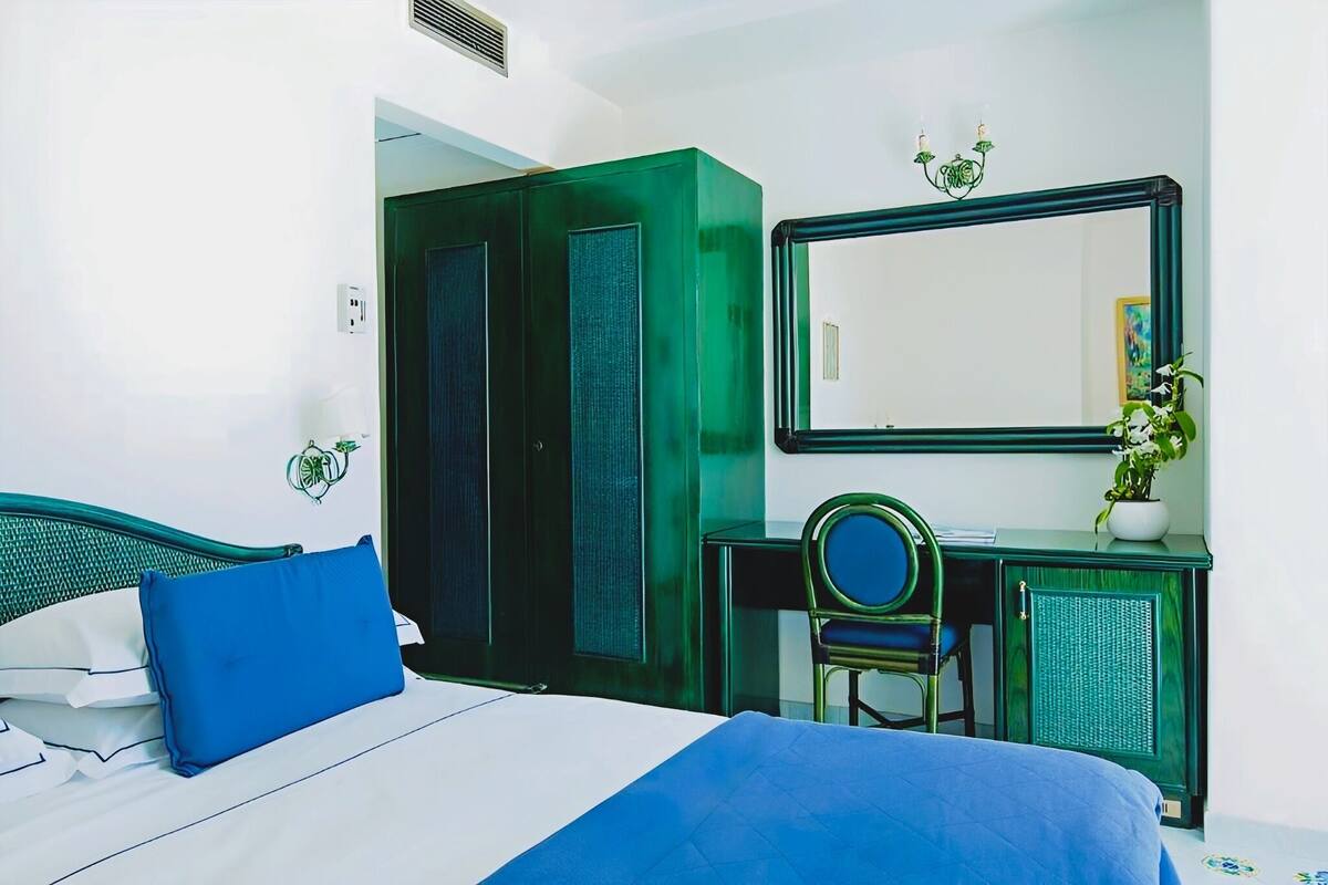 A hotel room with a bed, a green wardrobe, a desk with a chair, and a mirror.