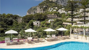 A swimming pool with lounge chairs and umbrellas, surrounded by greenery and buildings on a hillside. - Hotel La Vega (Capri)