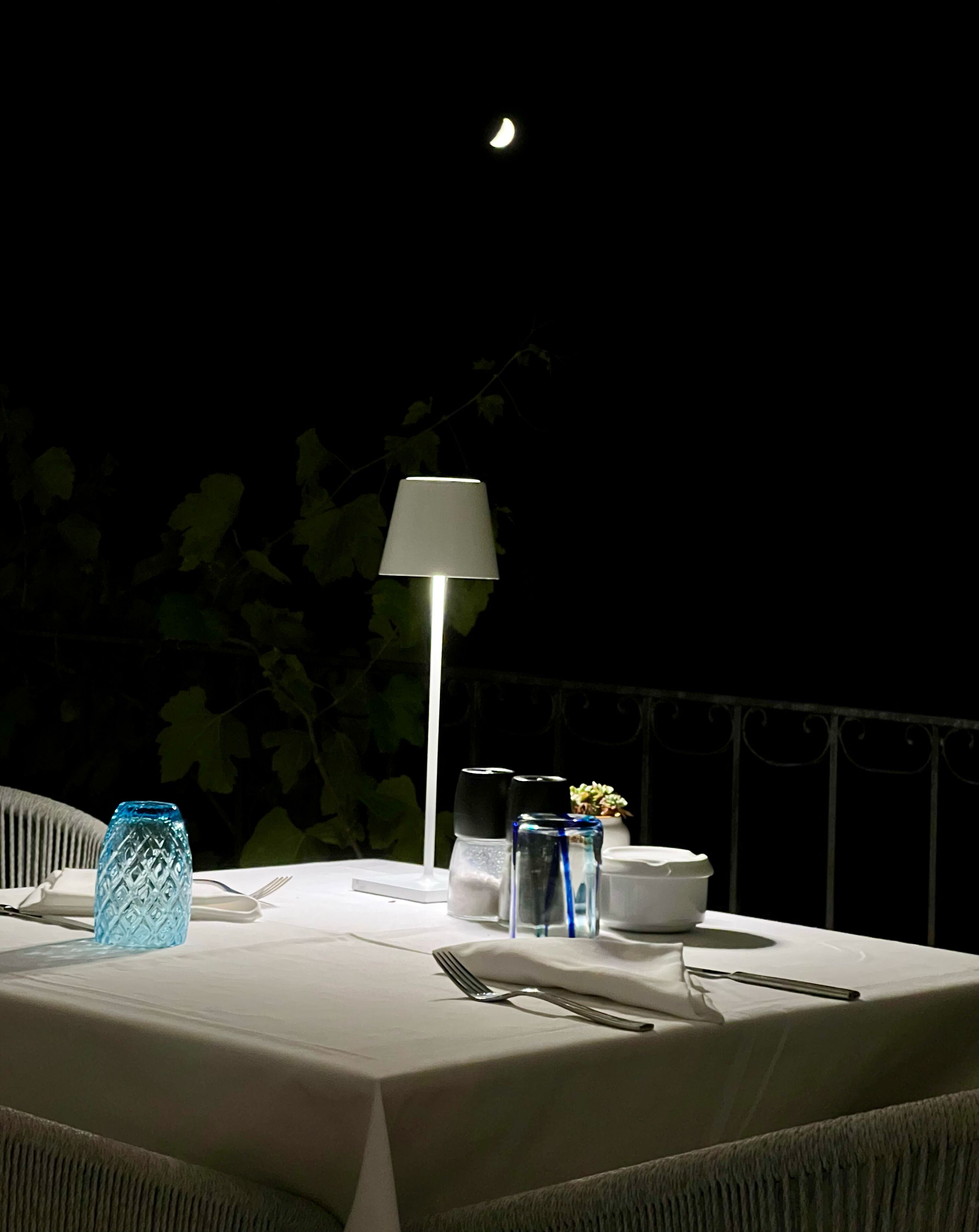 An outdoor dining table set for two with a white tablecloth, a blue glass vase, a white candle holder, and a white table lamp.