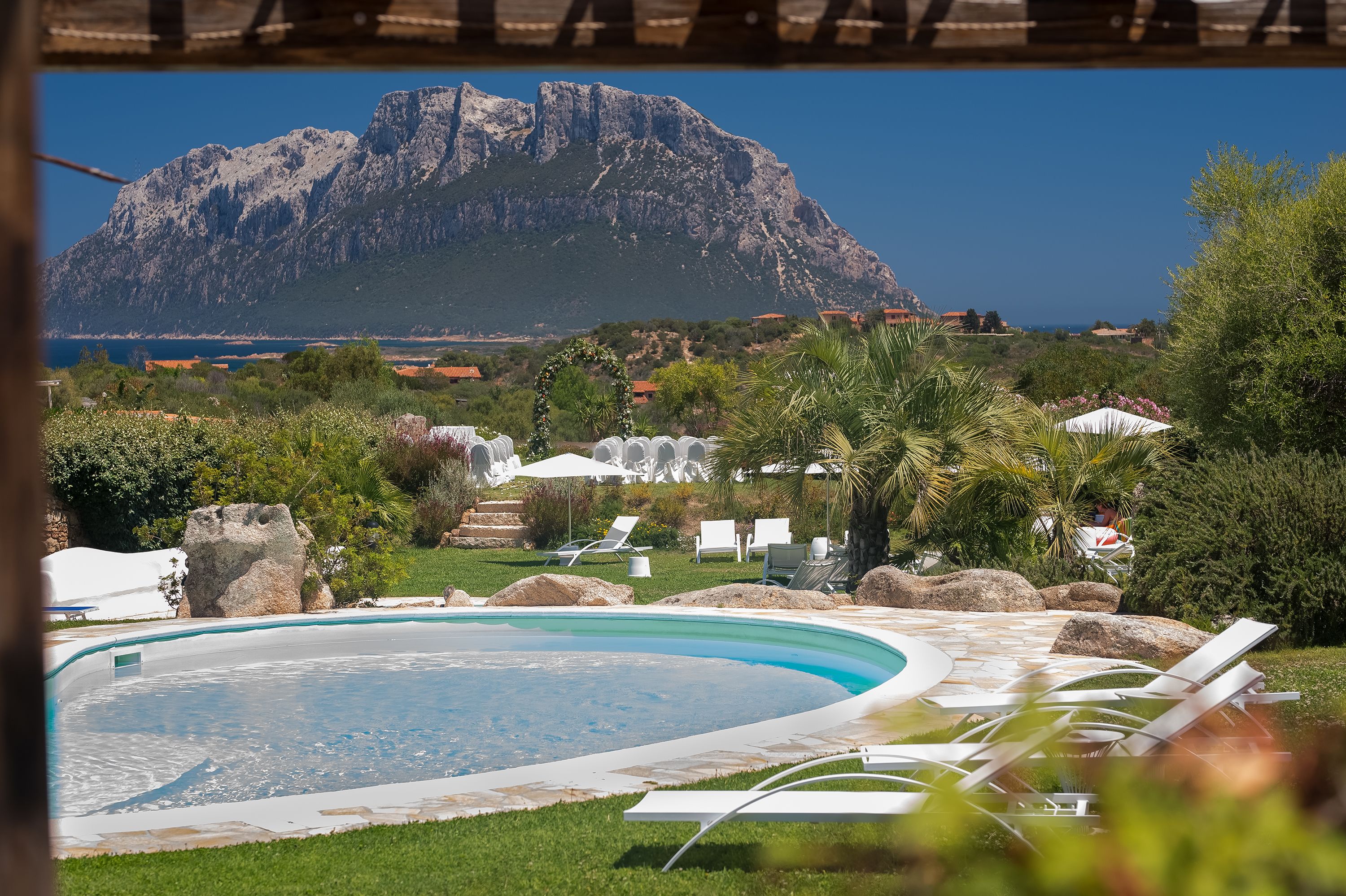A pool with lounge chairs, a rock formation, and a view of a mountain and coastline.
