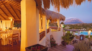 A dining area with a table set for two, a thatched roof, and a view of a mountain and pool in the background. - Hotel Ollastu (Olbia)