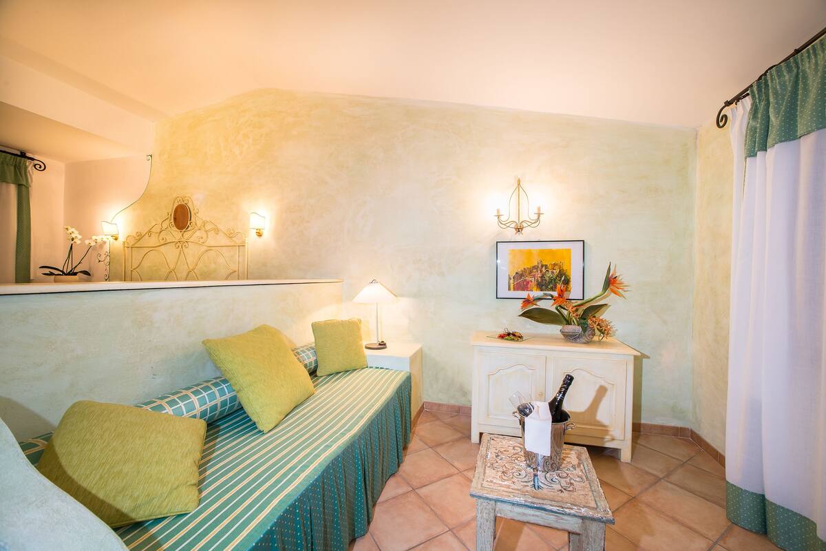 A cozy room with a bed, a small table with a bottle, and a wall-mounted shelf with decorative items.
