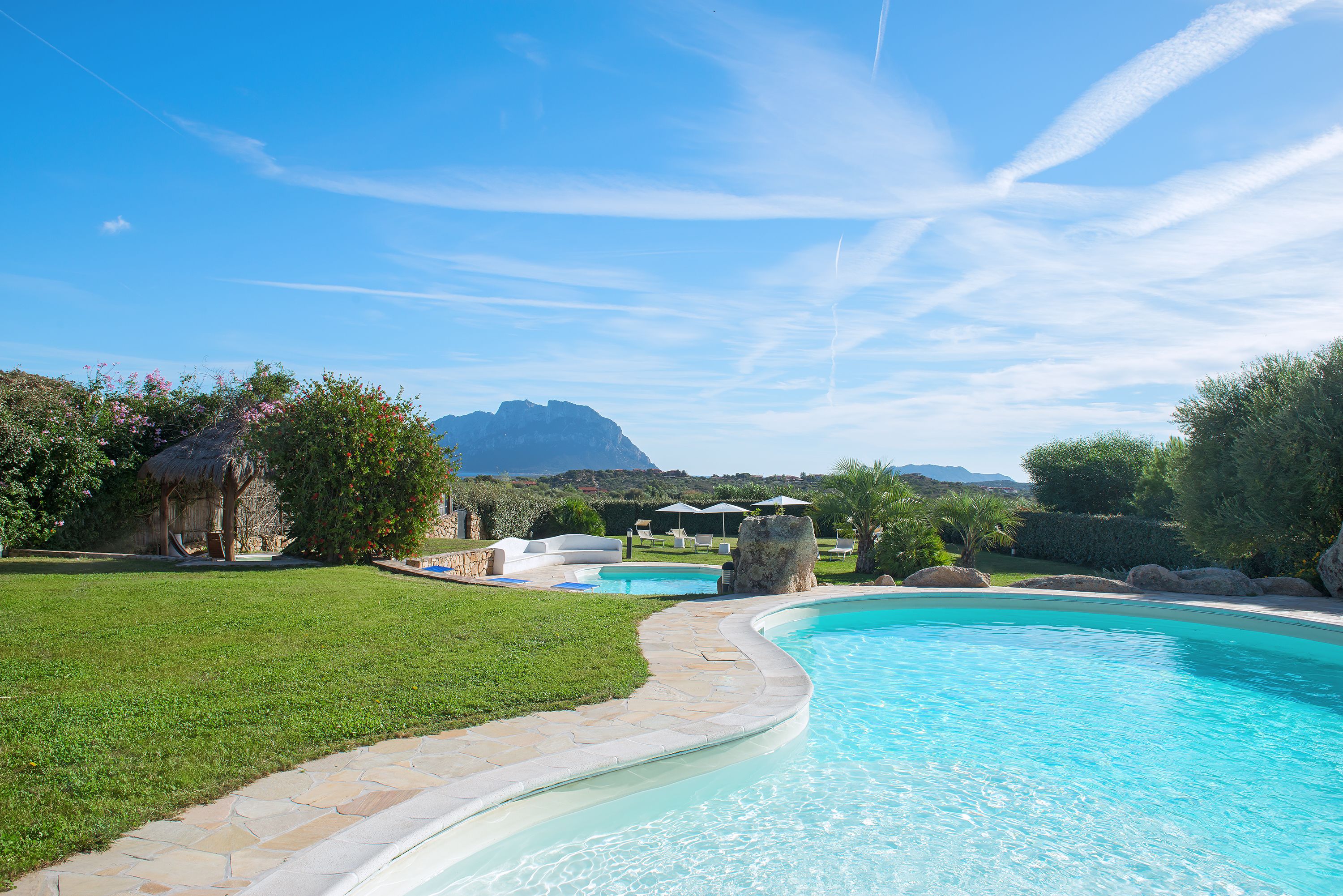 A swimming pool with a stone pathway, lounge chairs, and umbrellas.