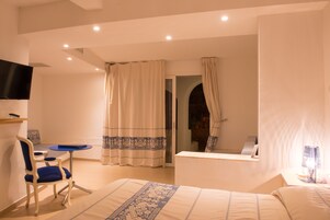 Habitación de hotel con cama, una silla, una mesita, televisor y baño con ducha y espejo.