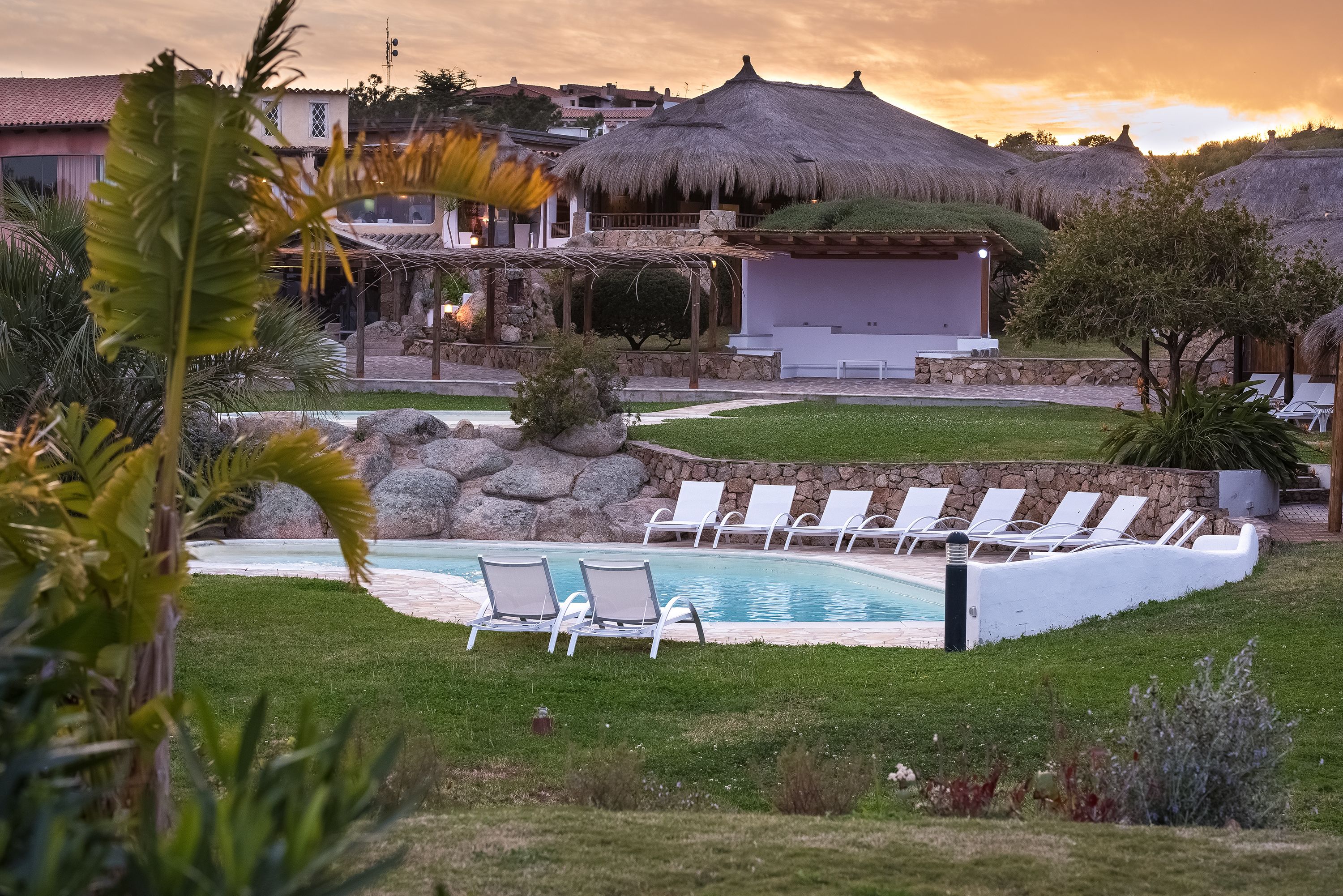 A pool area with lounge chairs, a thatched-roof building, and a well-maintained lawn.