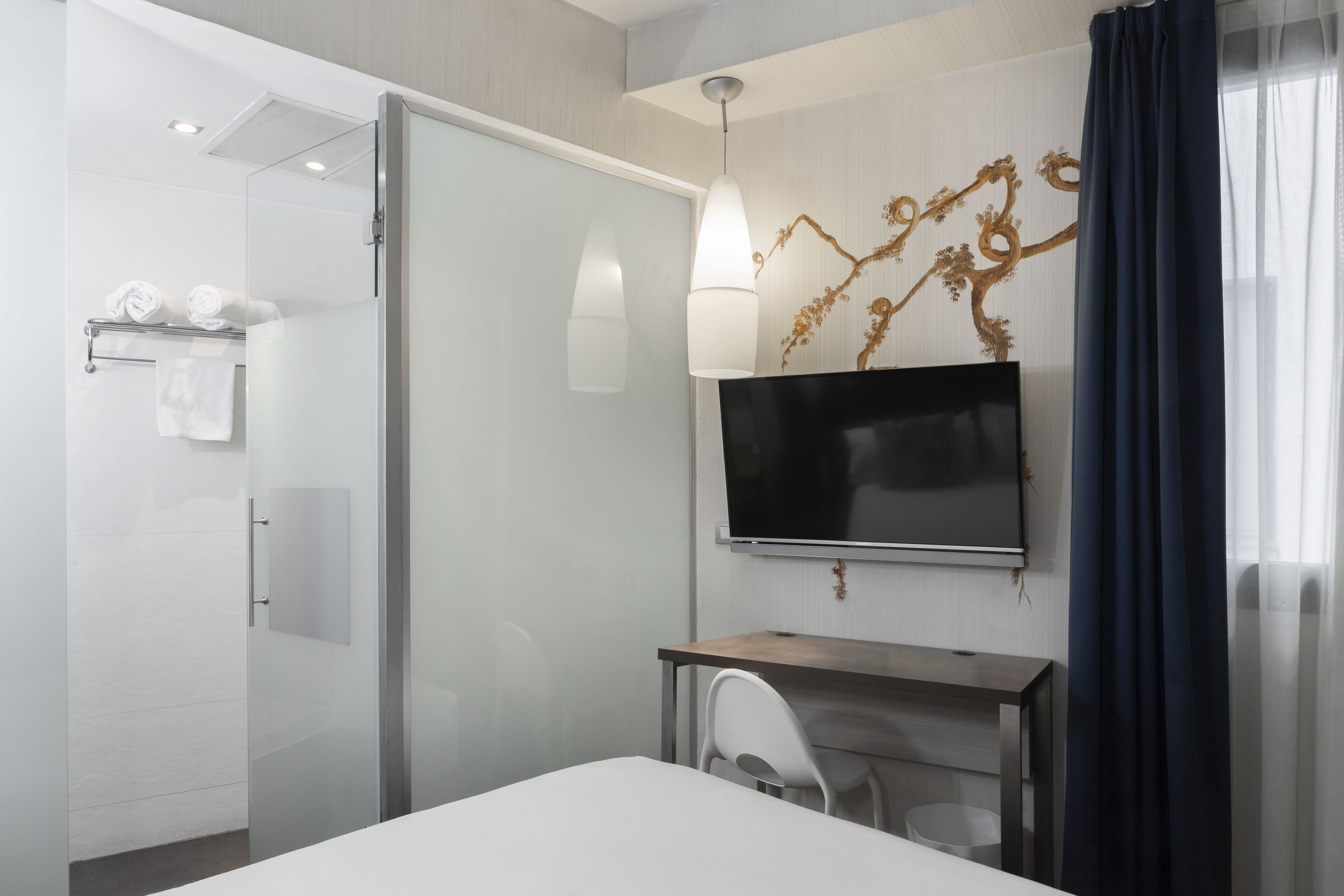 Una habitación de hotel moderna con cabina de ducha de vidrio, un televisor de pantalla plana montado en la pared, un escritorio de madera y una silla blanca.