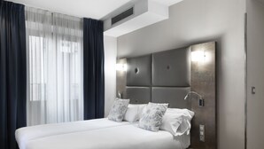 Una habitación de hotel moderna con una cama grande, un cabecero con iluminación integrada, una mesita de noche y una ventana con cortinas translúcidas.