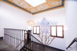 A staircase with a wooden handrail and black balusters, leading to a landing with a decorative ceiling and wall-mounted lights.