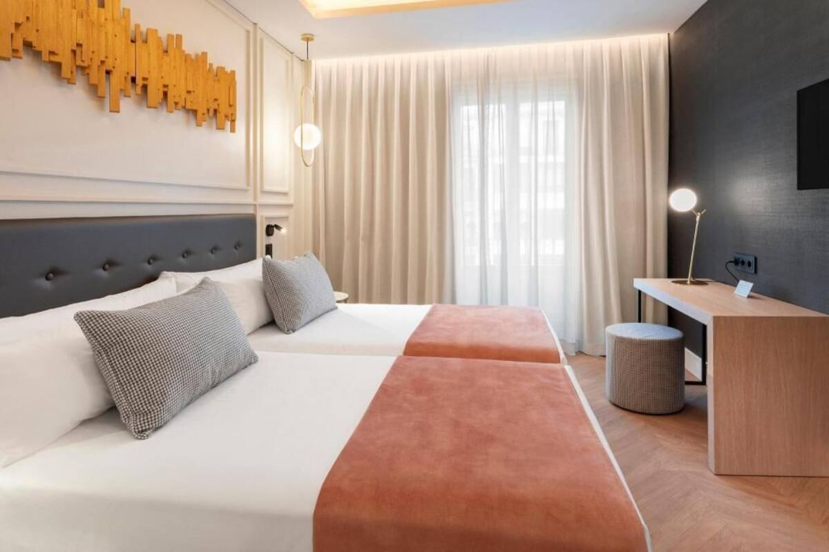 A hotel room with two beds, a desk, a lamp, and a large window with curtains.