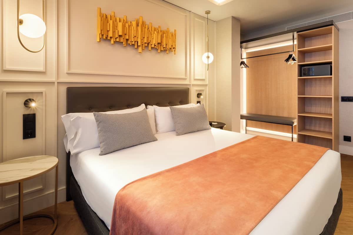 A modern hotel room with a large bed, a bedside table, a wall-mounted light, and a built-in wardrobe.