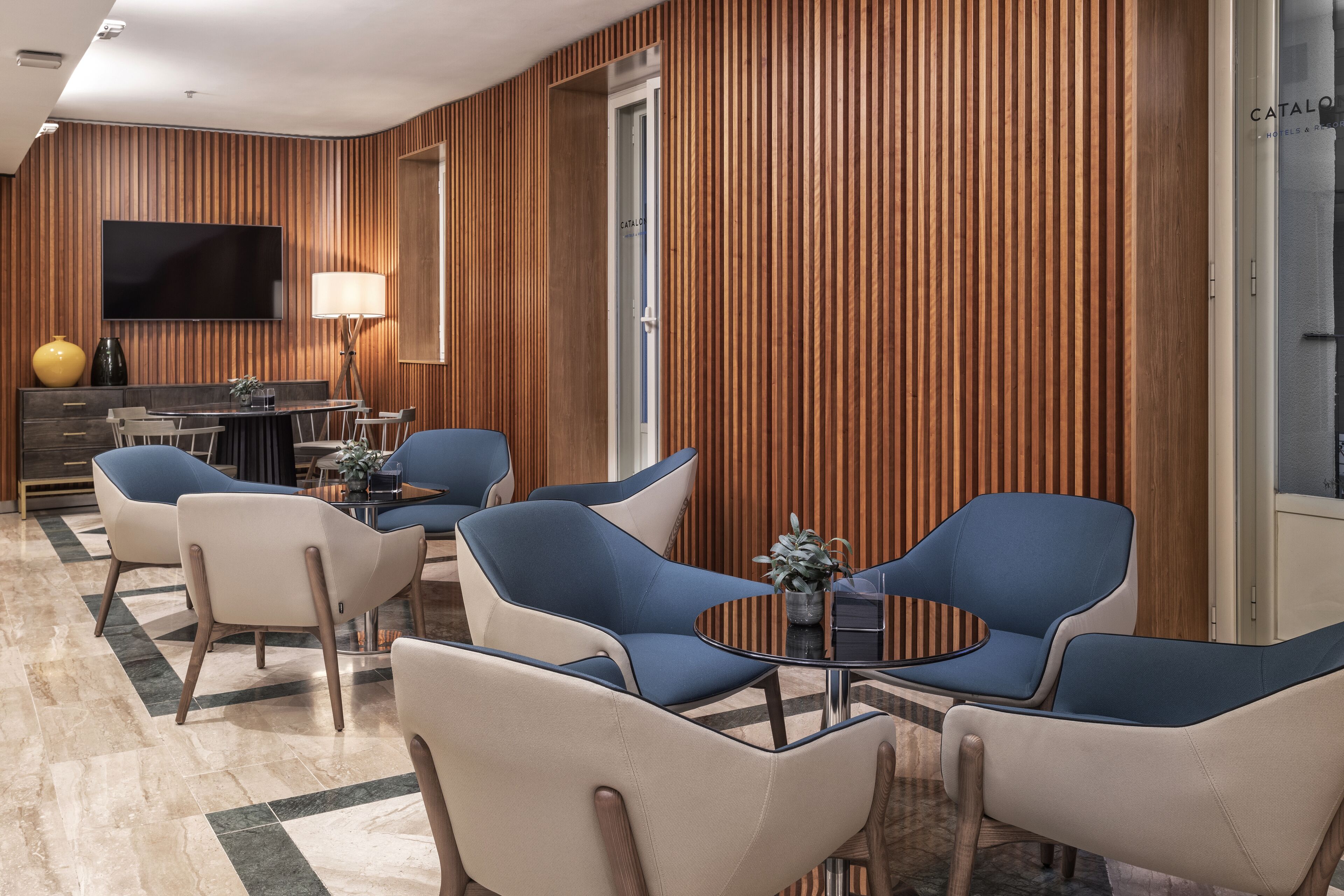 A modern hotel lobby with a wooden panel wall, a flat-screen TV, a dining area with blue and beige chairs, and a round table.