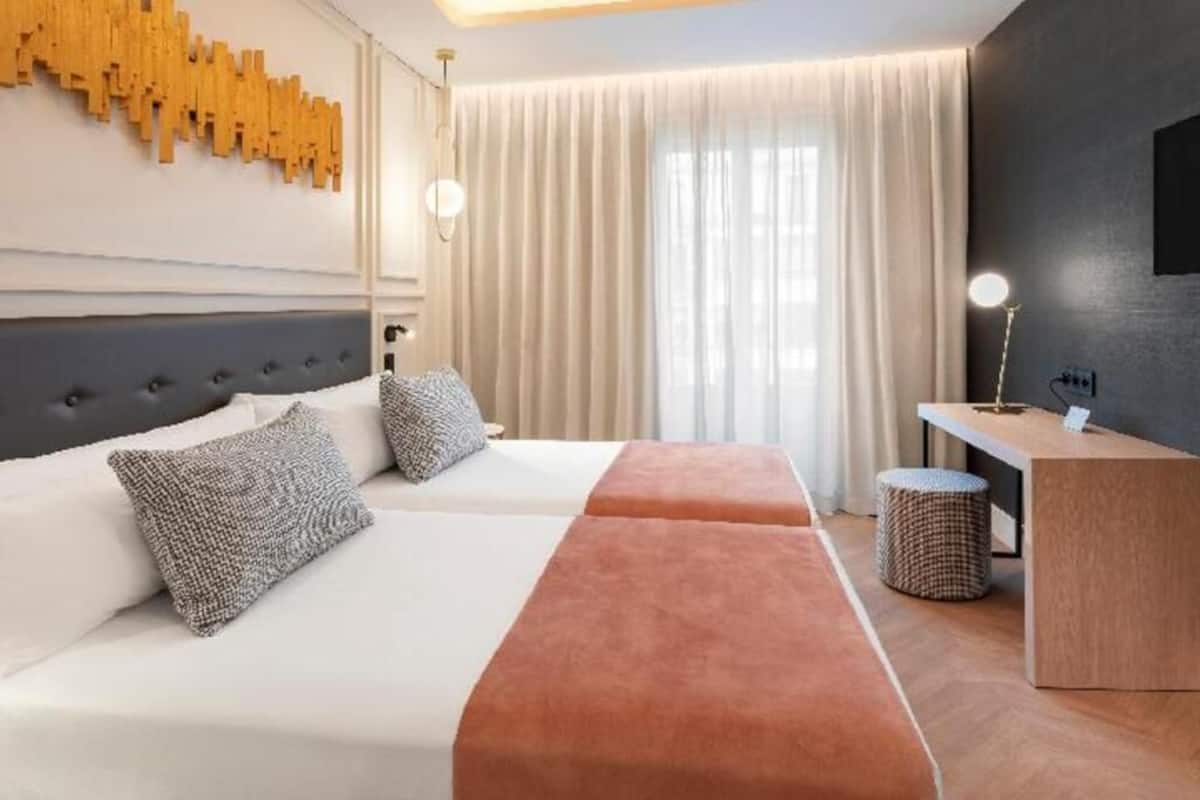 A hotel room with two beds, a desk, a lamp, and a large window with curtains.
