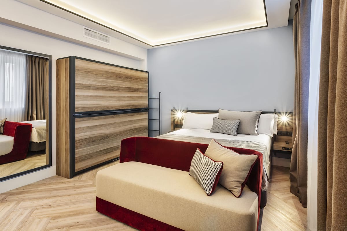A modern hotel room with a large bed, a red sofa, a wooden accent wall, and a mirror.
