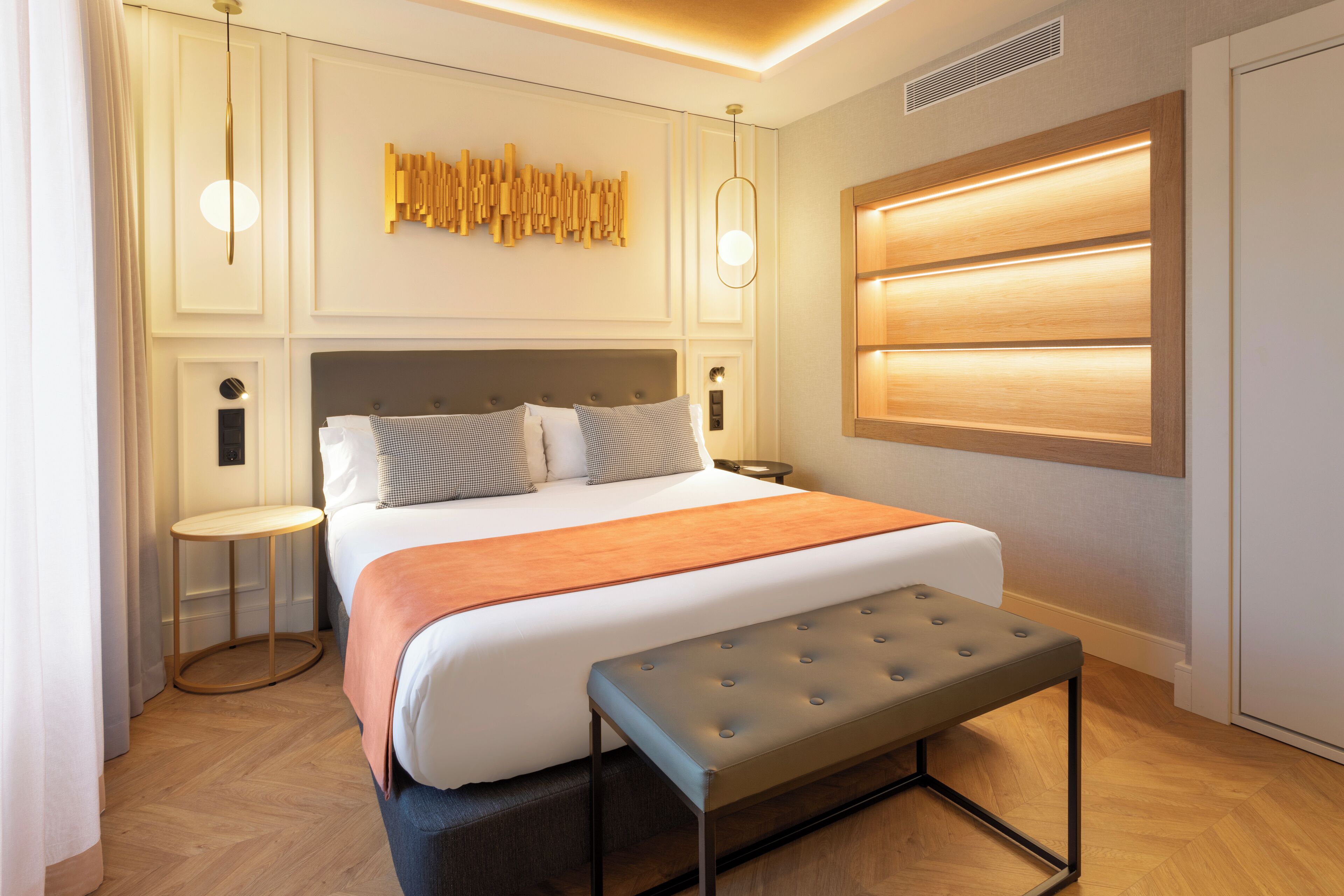 Une chambre d’hôtel moderne avec un grand lit, une banquette, une table de chevet, une lampe fixée au mur et un élément décoratif mural.
