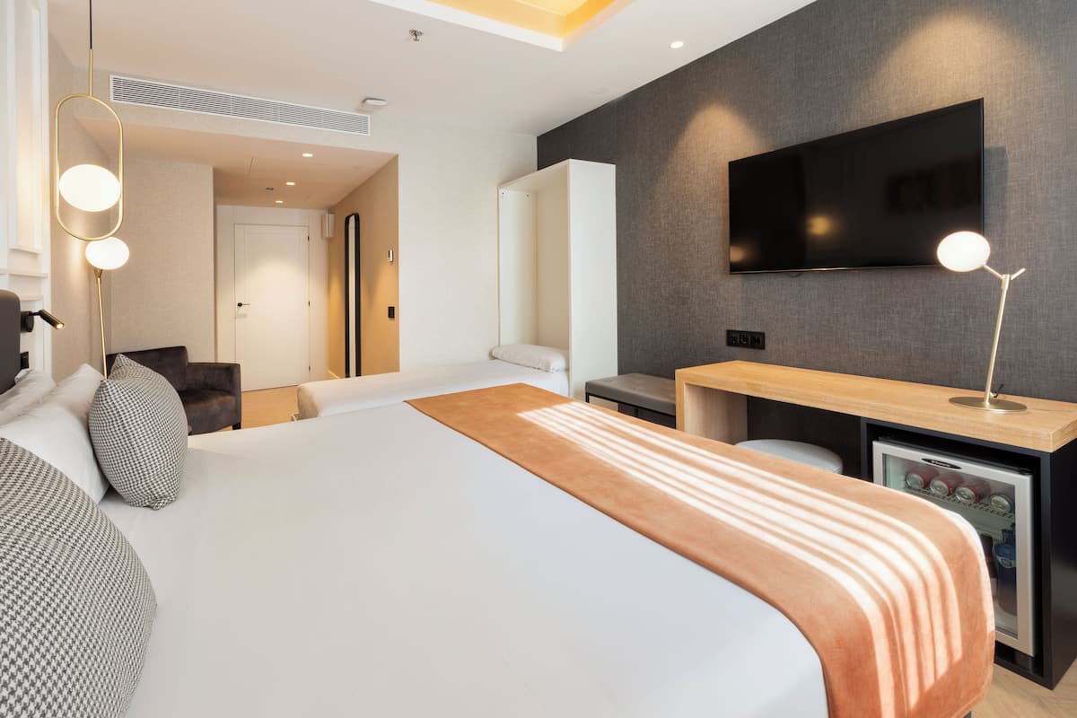 A modern hotel room with a large bed, a desk with a flat-screen TV, a minibar, and a seating area with a sofa and armchairs.