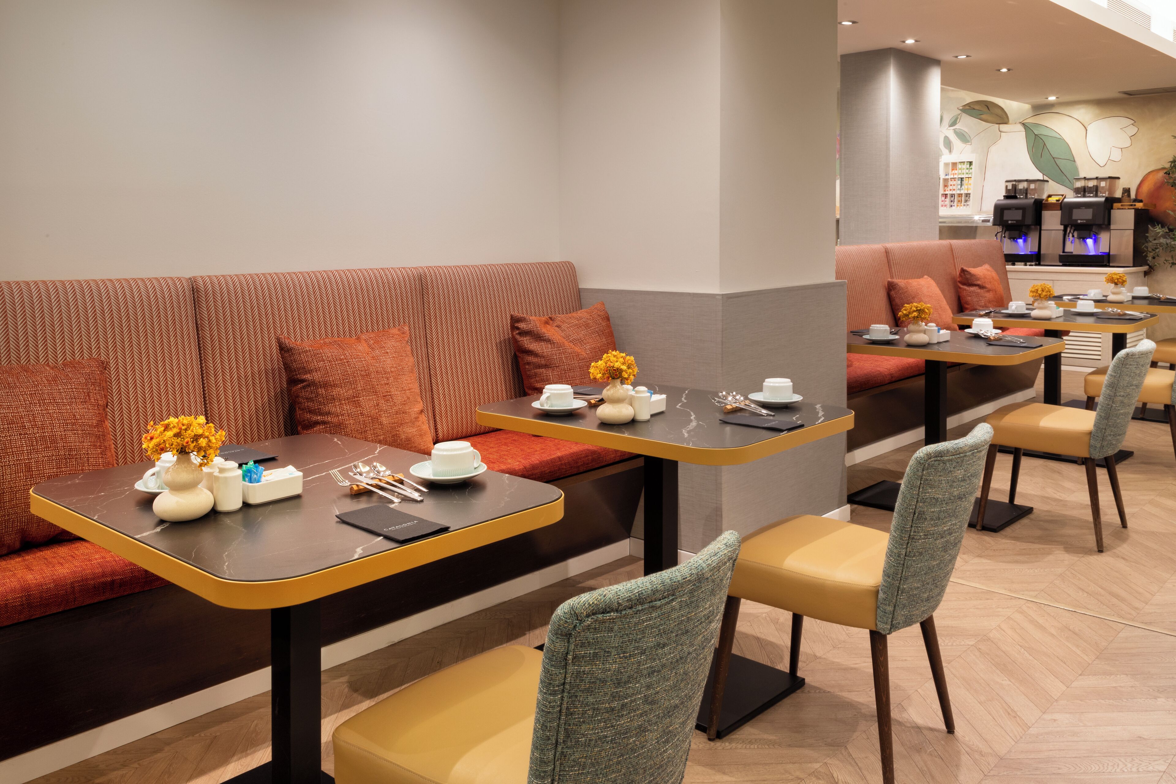A modern dining area with a long table, cushioned seating, and a coffee machine in the background.