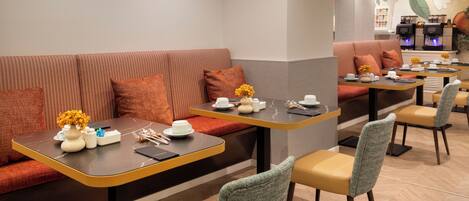 A modern dining area with a long table, cushioned seating, and a coffee machine in the background.