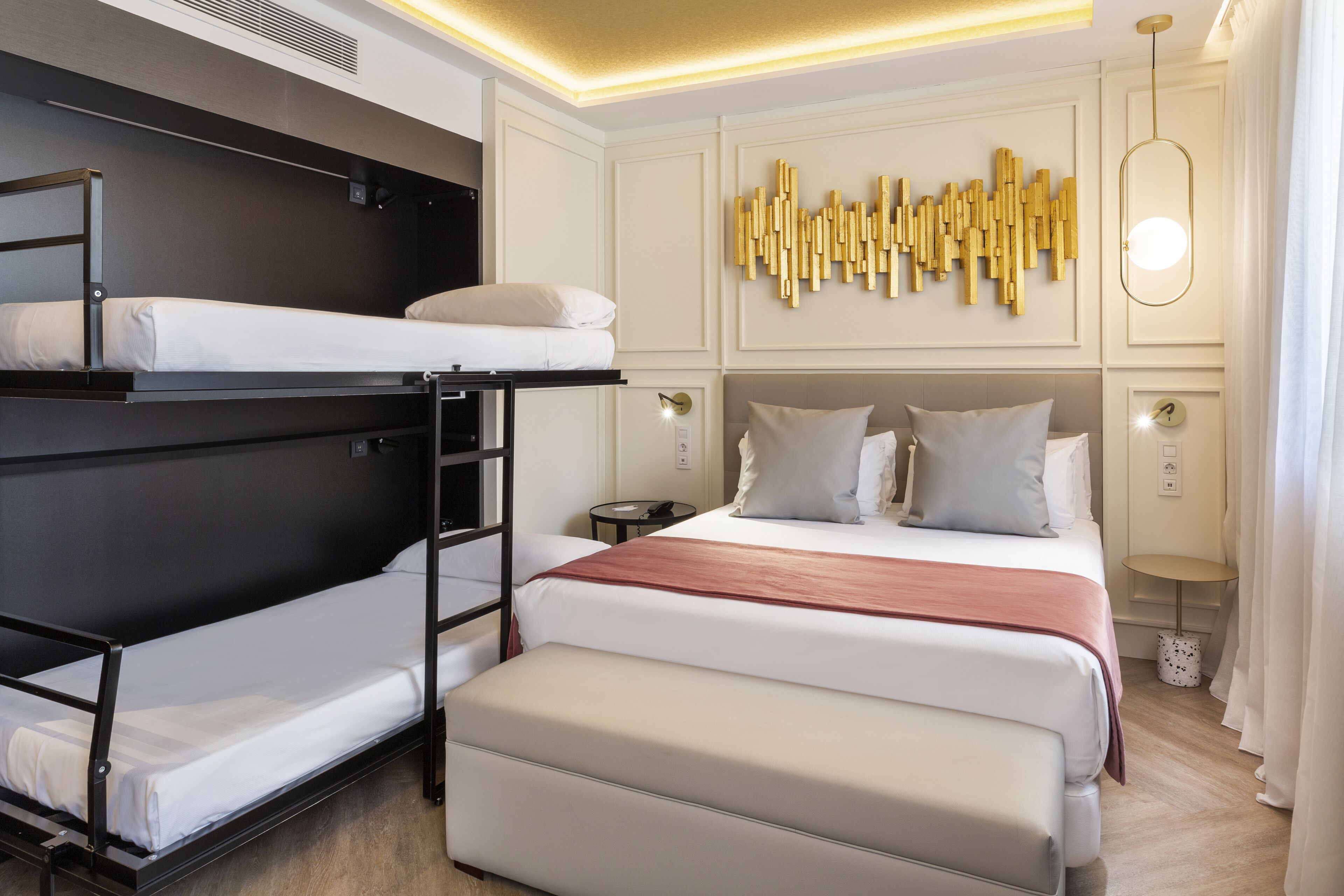 Habitación de hotel con literas, una cama grande, una mesita de noche, una lámpara de pared y un elemento decorativo en la pared.