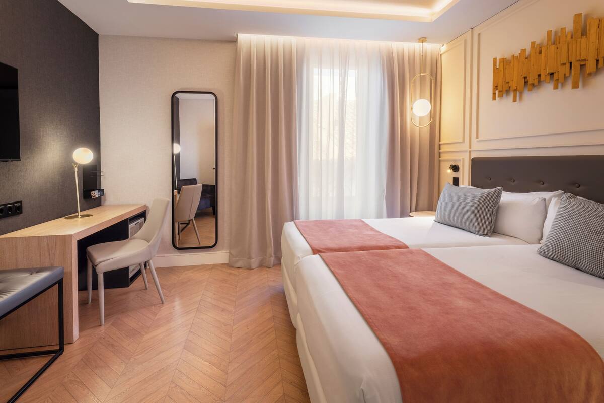 A hotel room with a bed, a desk, a chair, a mirror, and a large window with curtains.